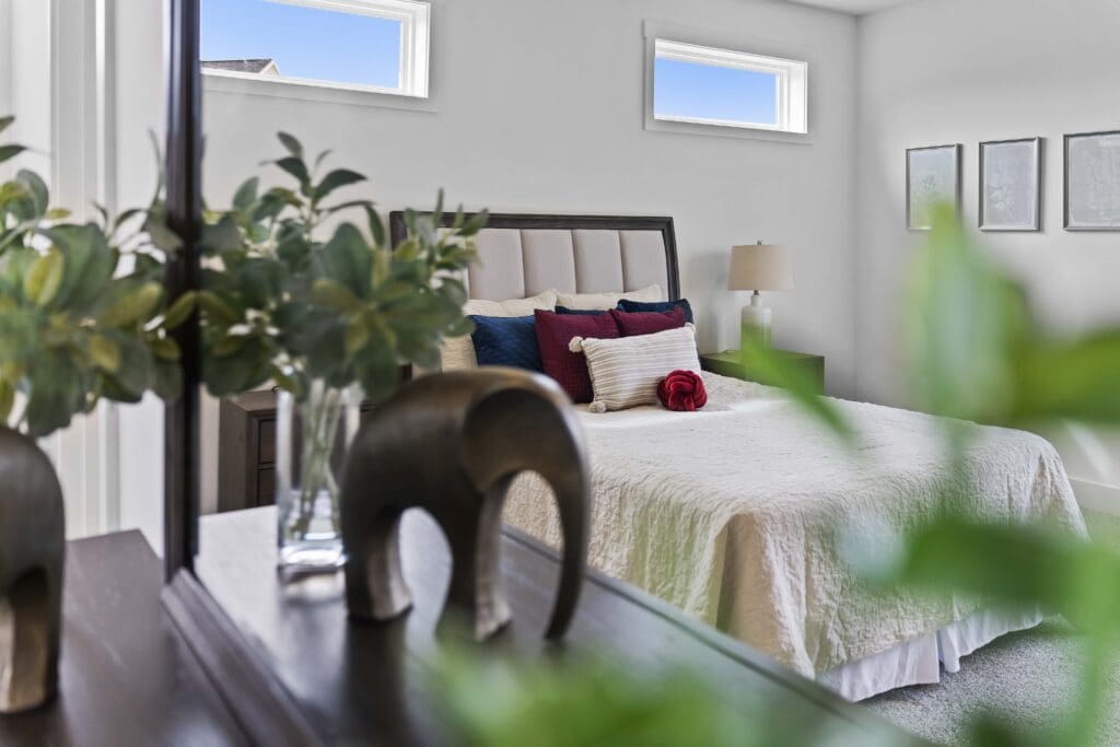 A neatly made bed with white bedding, accent pillows, and a red flower sits in a bright bedroom; perfect inspiration for home remodeling Harrisburg homeowners can trust. A dresser with a plant and elephant figurine is in the foreground.