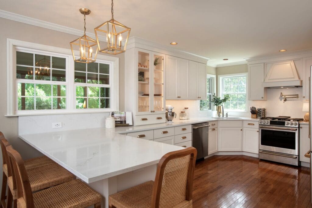 Modern kitchen with white cabinets, marble countertops, stainless steel appliances, wooden floor, and wicker barstools, beautifully lit by pendant lights and natural light—perfect inspiration for Central PA home renovations.