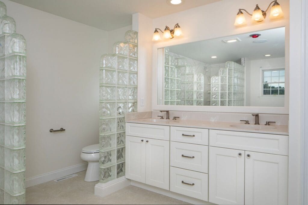 Modern bathroom with double sink vanity, large mirror, glass block walls, white cabinets, and a toilet separated by a glass block partition—perfect inspiration for your next home remodeling Harrisburg project.
