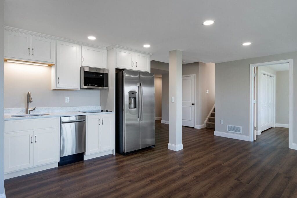 Basement remodel that includes a kitchenette with white cabinets, stainless steel appliances, wood flooring, and an open area leading to a staircase and doorway—perfect for those considering home remodeling Harrisburg with McNaughton Homes.