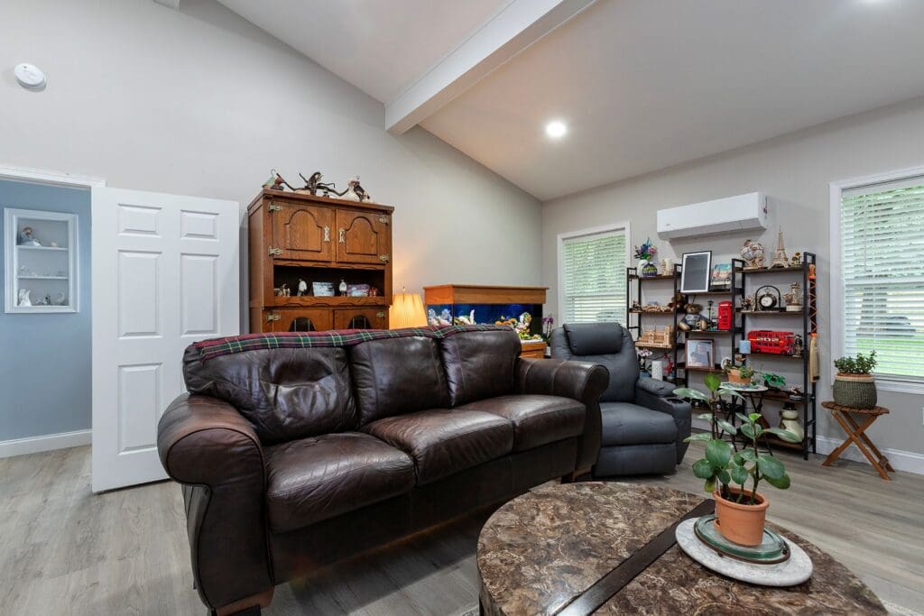 This home addition by McNaughton Homes features a brown leather sofa, recliner, wooden cabinet, and shelves with decor. A coffee table with a potted plant sits in the foreground, with windows brightening this inviting home remodeling Central PA space.