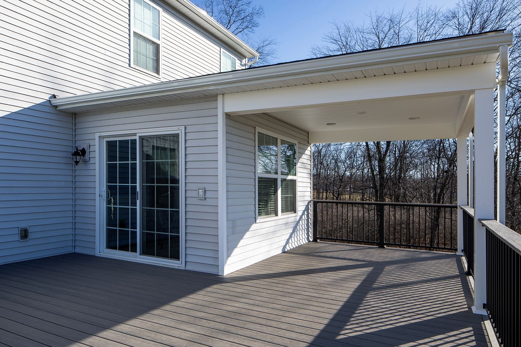 A covered residential deck with wood-like flooring, black railing, and sliding glass doors enhances this white house with siding—perfect inspiration for home remodeling in Harrisburg or Mechanicsburg. Bare trees are visible in the background.