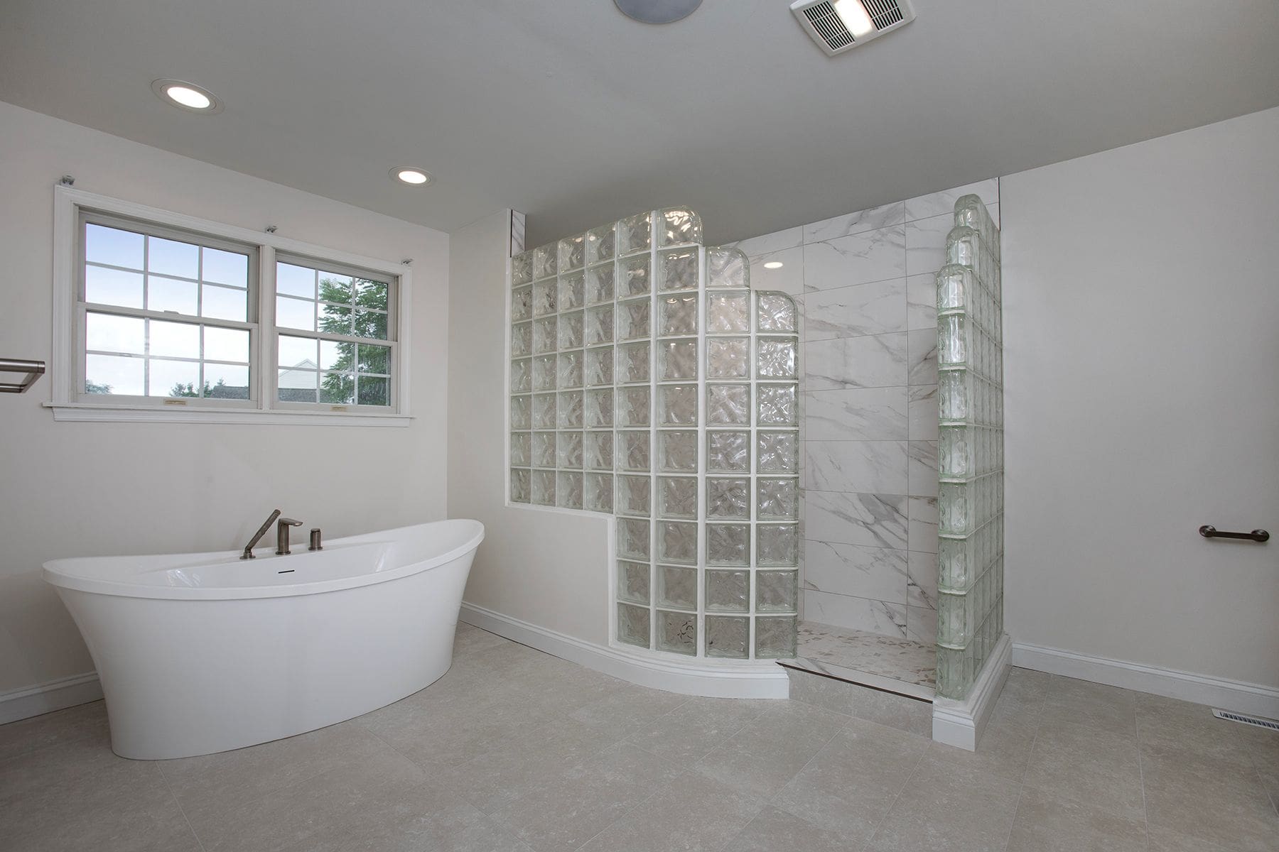 Modern bathroom with a freestanding bathtub, walk-in shower with glass block walls, marble tiles, and a large window—perfect inspiration for your home remodeling Harrisburg project.