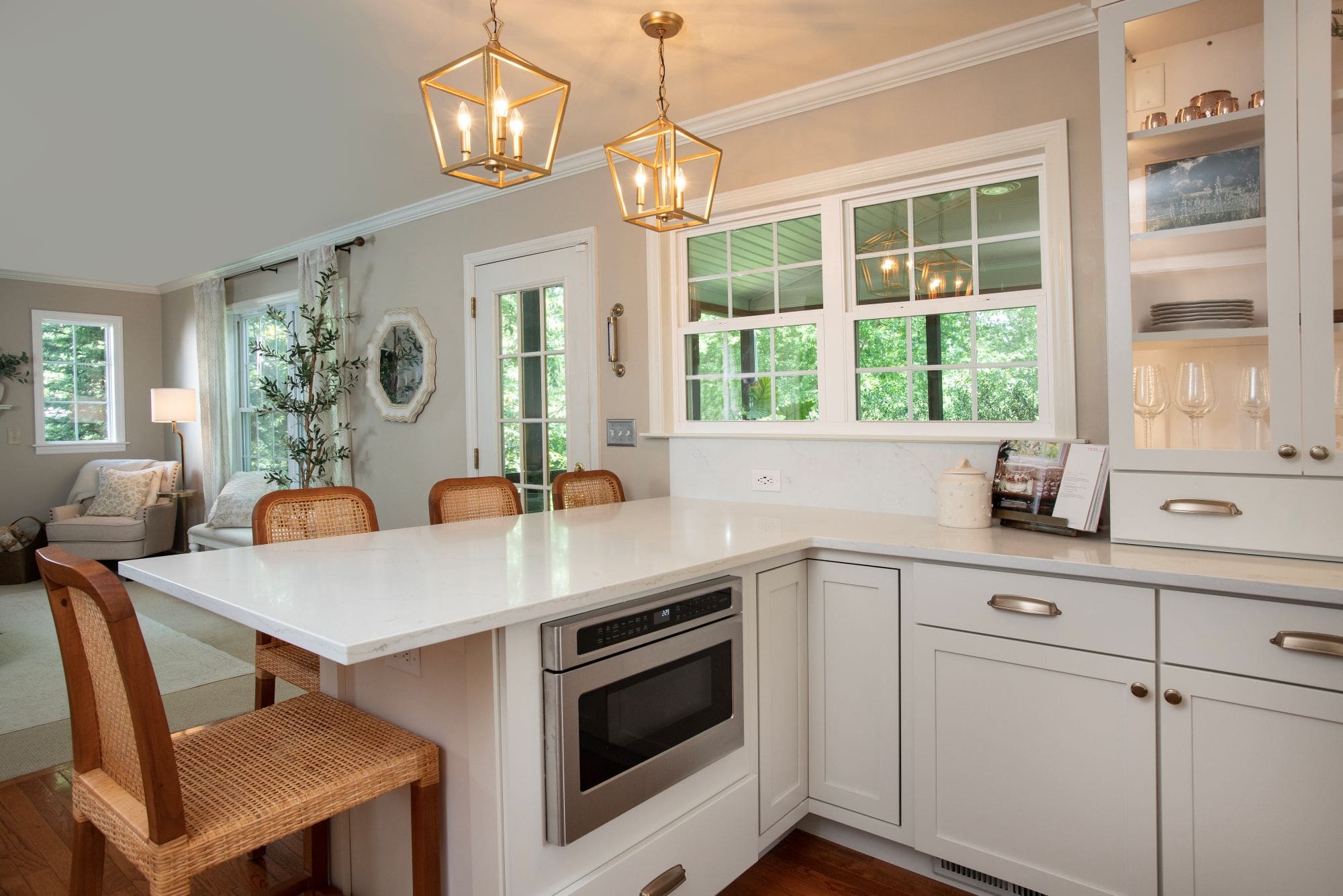 A modern kitchen with white cabinets, a built-in microwave, a white countertop, wicker chairs, pendant lights, and windows showing greenery—a perfect example of stylish kitchen remodeling in Harrisburg.