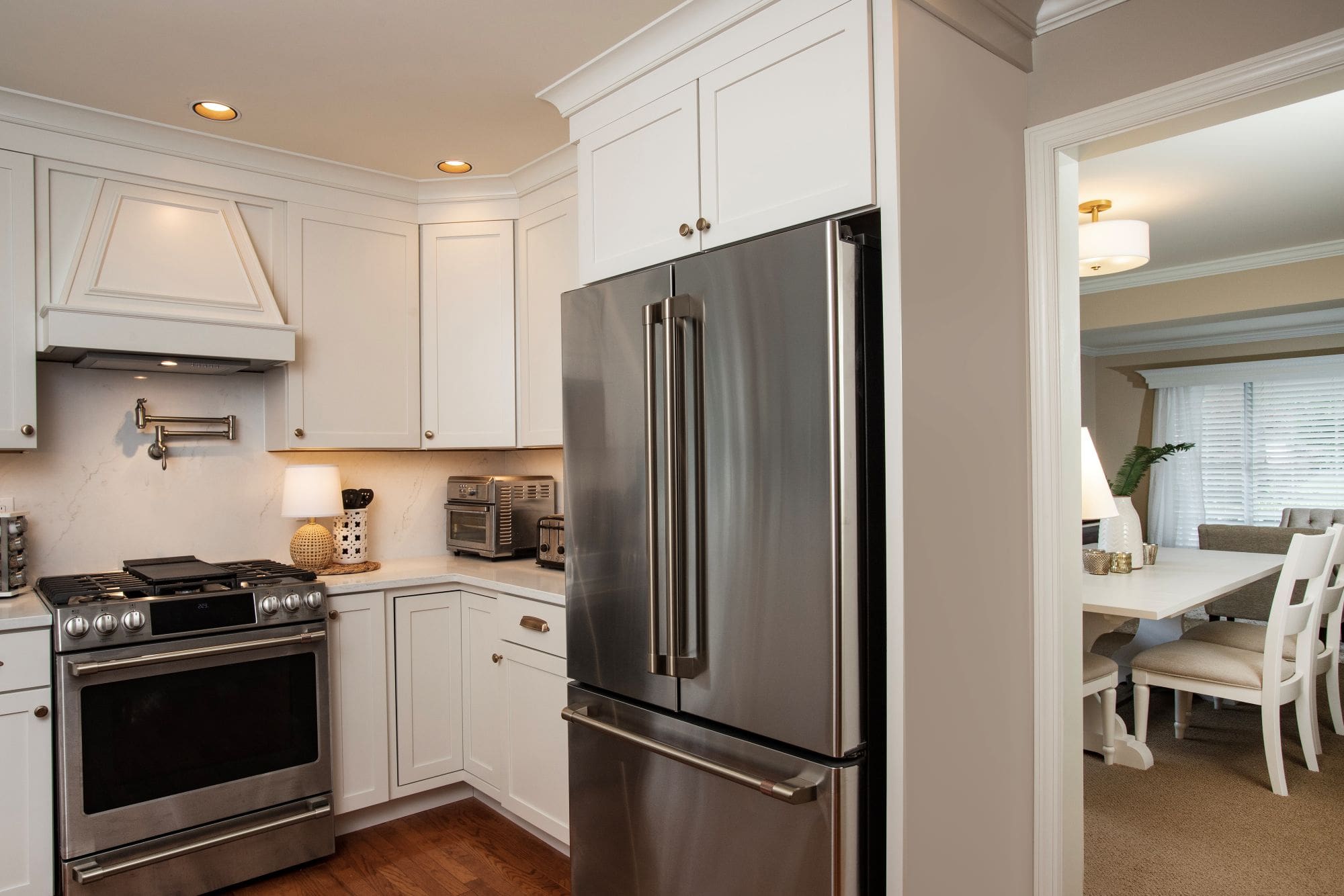 Modern kitchen with white cabinets, stainless steel appliances, wood flooring, and an open area leading to a staircase and doorway—perfect for those considering home remodeling Harrisburg with McNaughton Homes.